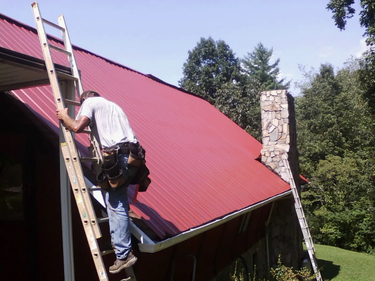Roofer on a ladder beside a red metal roof and stone chimney — Soffit & Fascia Repair by Roofing Brothers in Ebensburg